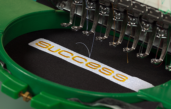 An embroidery machine financed by Beacon Funding stitches the word "Success" into black fabric.