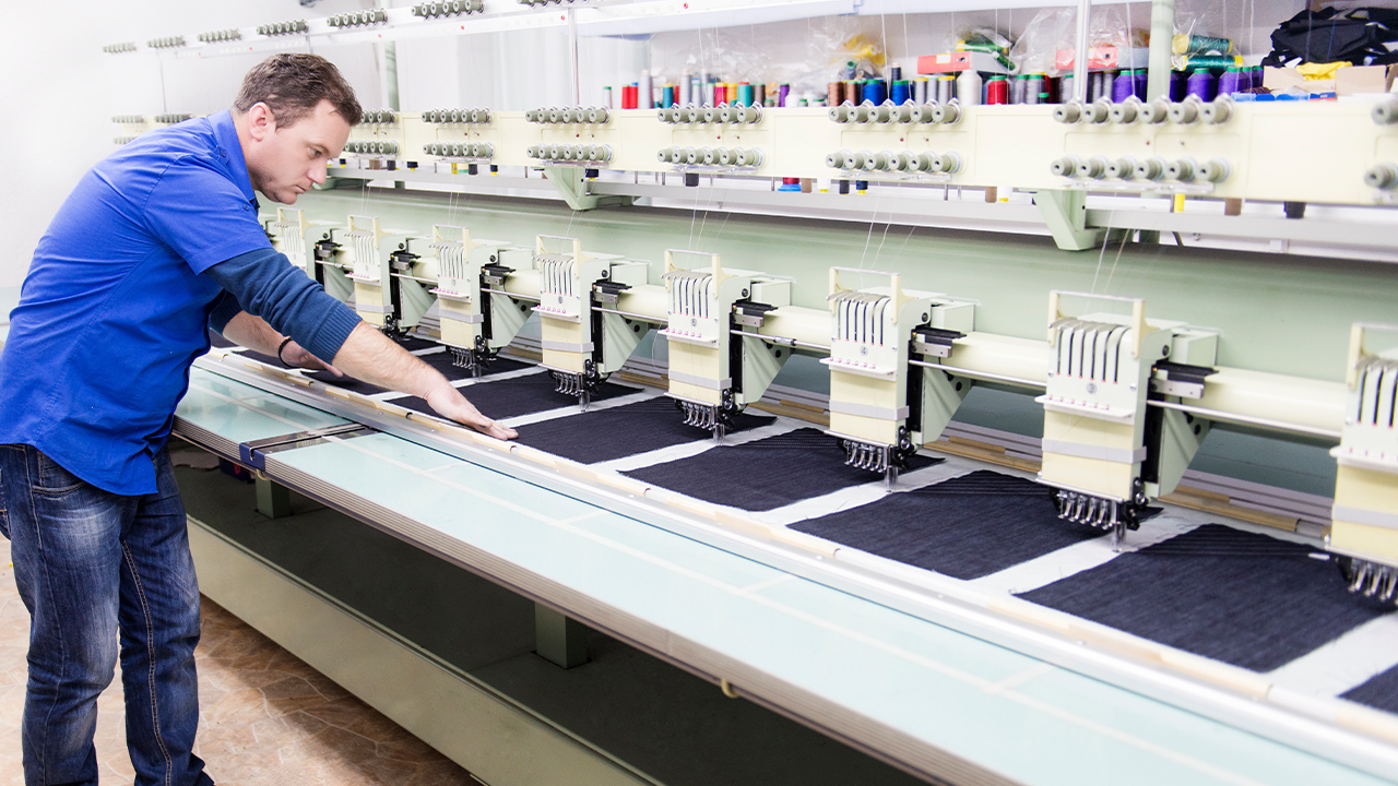 A business owner wearing a blue shirt inspects his multi-head embroidery machine.