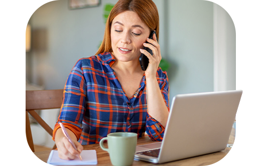 A woman in a plaid shirt is on the phone while writing notes at a table with a laptop and a cup of coffee.