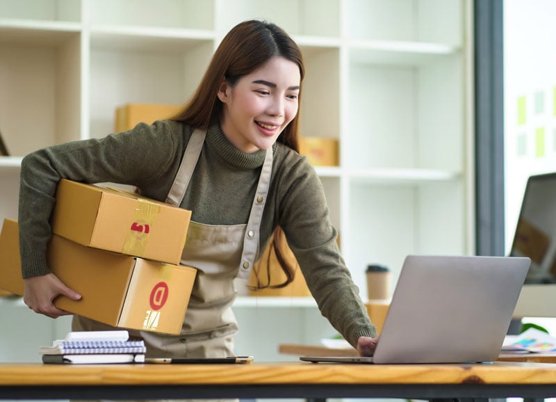 Woman on computer smiling holding boxes.