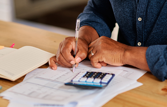 Closeup of business owner penciling in their tax benefits on paper after using a blue handheld calculator on their wooden desk.