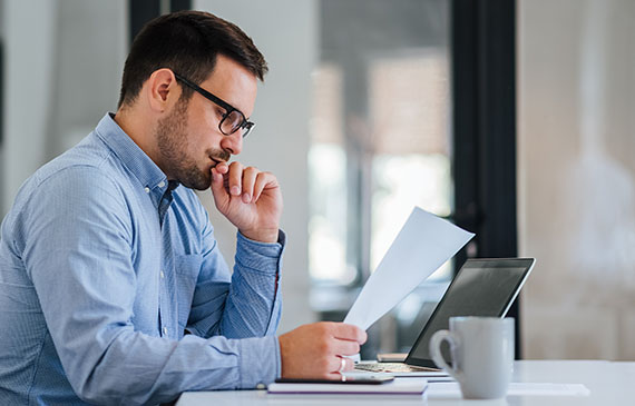 Business owner wearing glasses and a blue buttoned-up shirt reviews his monthly expenses on a sheet of paper.