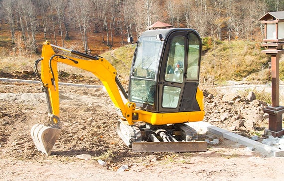 A bright-yellow mini-excavator sits in the middle of a landscaper's dig site. A bright-yellow mini-excavator sits in the middle of a landscaper's dig site.