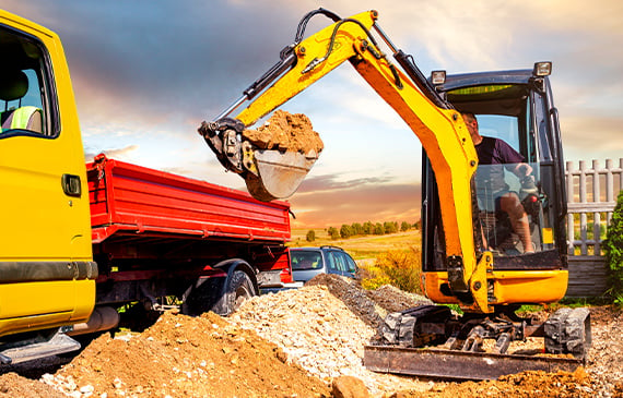 A landscaper operating a yellow mini excavator unloads a scoop of dirt onto a dump truck.