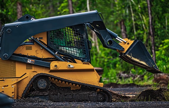 A landscaper drives a yellow compact loaders and begins lowering his bucket to flatten a layer of soil. A landscaper drives a yellow compact loaders and begins lowering his bucket to flatten a layer of soil.