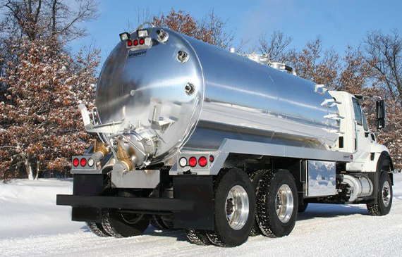 A large white vacuum septic truck drives backwards in the middle of winter with snow surrounding a wooden landscape.