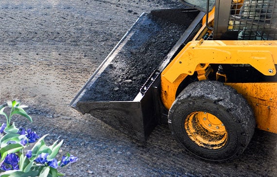 A yellow skid steer hauls a bucket of dirt over a ground plot.