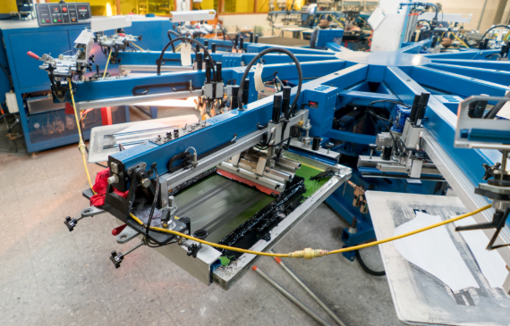 A closeup view of a blue, large automatic screen printing machine in the middle of pressing a garment. A closeup view of a blue, large automatic screen printing machine in the middle of pressing a garment.