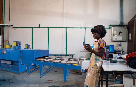 A business owner checks her mobile phone while standing in front of a blue electric dryer as t-shirts flow through its conveyer belt. A business owner checks her mobile phone while standing in front of a blue electric dryer as t-shirts flow through its conveyer belt.