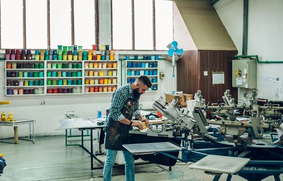 A screen printing operator presses a t-shirt using an automatic screen printing machine with many colored inks stacked on an organized wall behind him. A screen printing operator presses a t-shirt using an automatic screen printing machine with many colored inks stacked on an organized wall behind him.