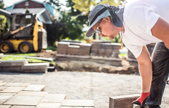 Hardscaping professional installing pavers with heavy equipment in the background, illustrating how Beacon Funding’s equipment financing helps contractors afford the machinery they need to take on more landscaping projects.