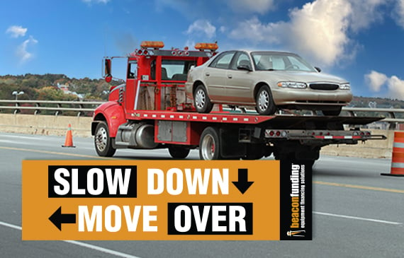 A red flatbed truck drives down the highway with a grey 4-door car on the back of the tow truck with the words "SLOW DOWN MOVE OVER" in the foreground.