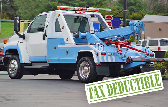 A white and blue tow truck sits in a parking lot with the words "TAX DEDUCTIBLE" underneath in green letters.