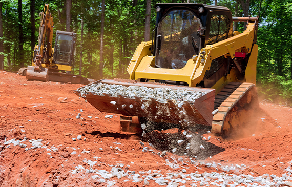 A compact track loader operator uses a bucket attachment to excavate a group of rocks for their hardscaping site.